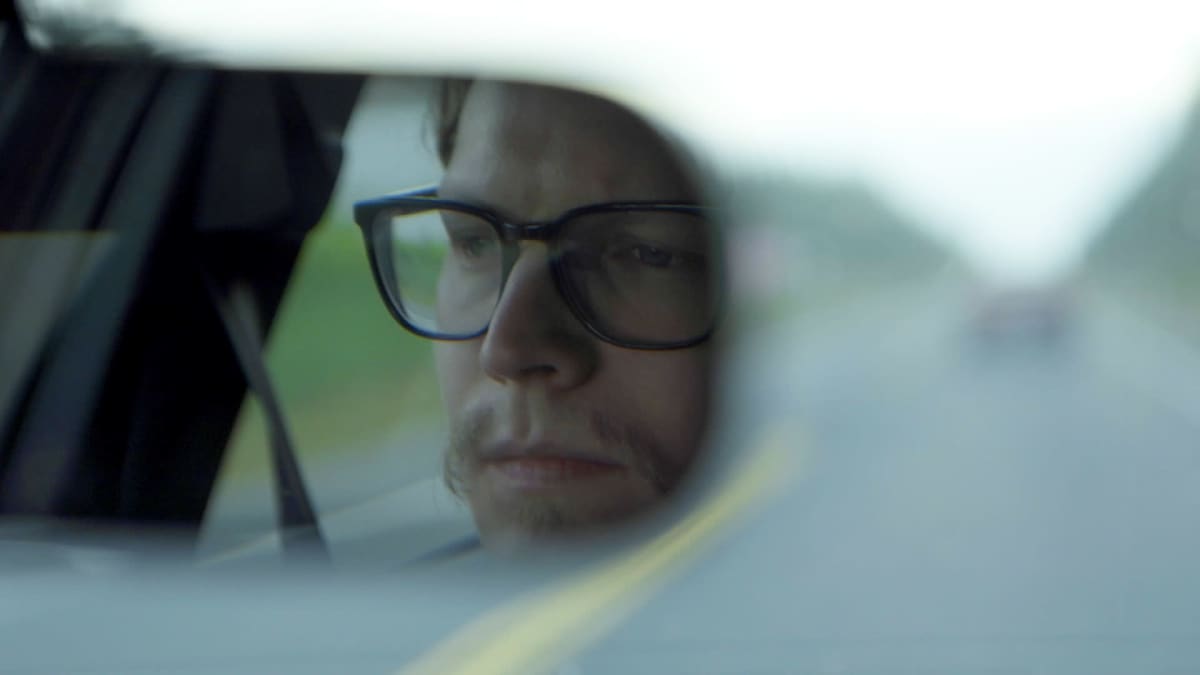 Rasmus ajaa autoa. Näkymä peruutuspeilistä Rasmuksen kasvoihin.