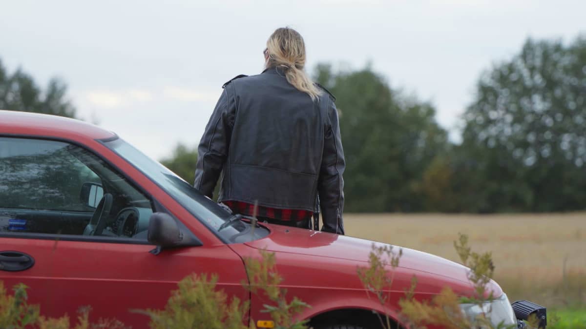 Rasmus istuu selkä kameraa kohti autonsa konepellillä ja katselee pohjalaista peltomaisemaa.