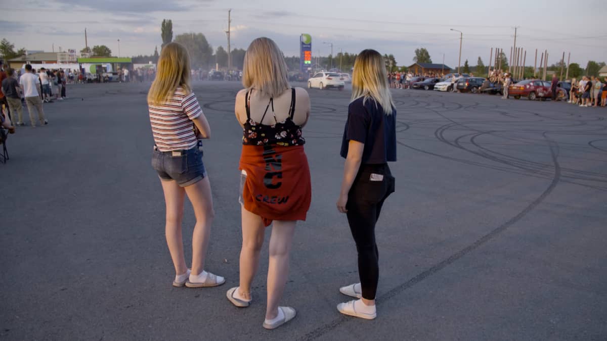 Kolme naista katselee, kun valkoinen auto polttaa kumia. 