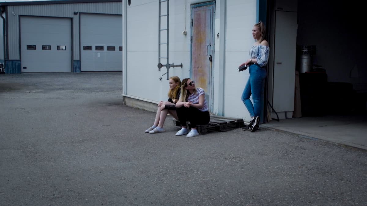 Kaksi naista istuu ja yksi seisoo autokorjaamon edustalla.
