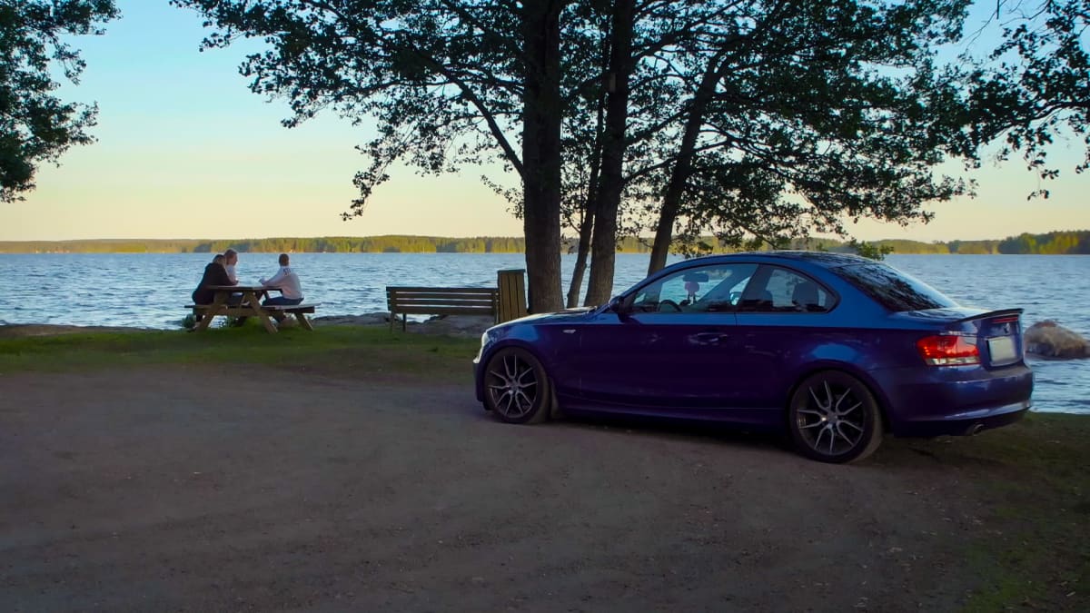 Kolme henkilöä istuu järven rannalla. Heidän vieressään on sininen auto parkissa.