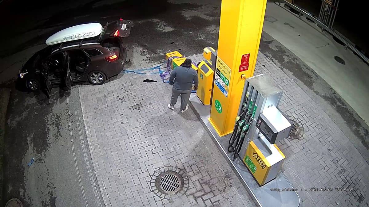 Valvontakamerakuva ylhäältäpäin,auto on kiinnitetty liinalla kiinni kaatuneeseen bensa-aseman maksuautomaattiin. Yksi mies seisoo selin kameraan asemalla.