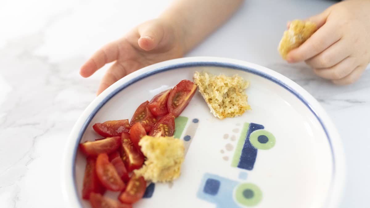 Pienen lapsen käsi ottaa lautaselta minitomaatteja ja leipäpalasia.