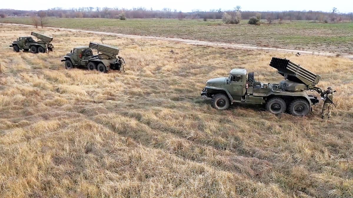 Pellolla on kolme kuorma-autoa, joissa on rakettipatteristot. Kunkin auton vieressä seisoo sotilas maastopuvussa.