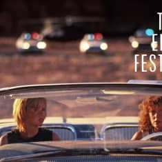 Thelma ja Louise istuvat autossa, takana näkyy poliisiautojen rivi. Kuva elokuvasta.