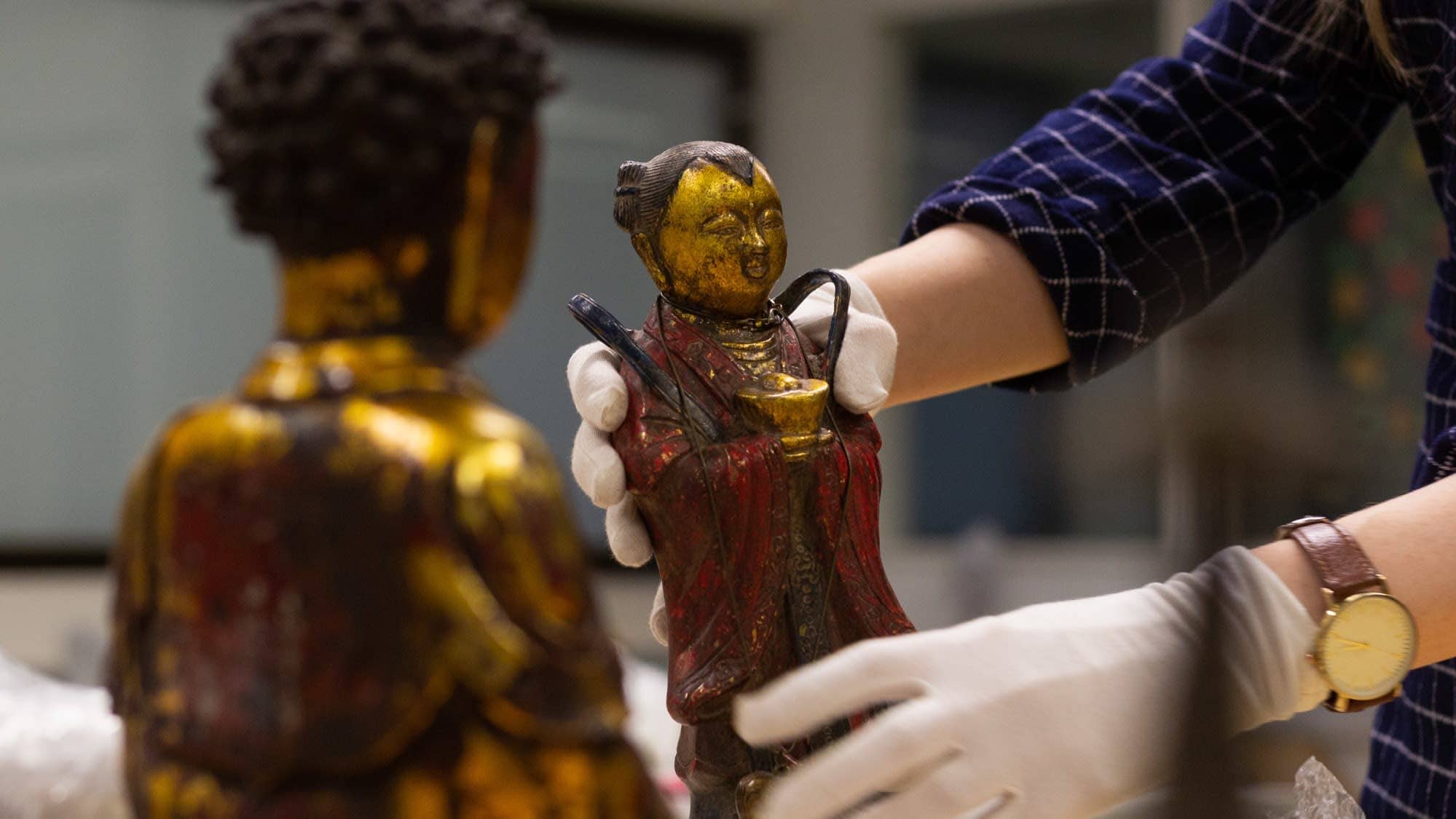 Aasialaisia taide-esineitä. Museon työntekijä nostaa yhtä pientä patsasta hansikkaat kädessä.