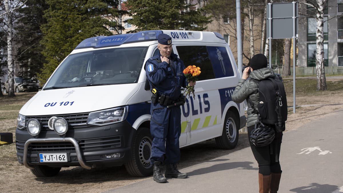 Nainen kuvaa kukkakimppu sylissä autoa vasten poseeraavaa poliisimiestä.