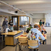 Oppilaita pulpeteissa syömässä kotitaloustunnilla tekemäänsä ruokaa. Kotitalousopettaja Elina Harju ja koulunkäyntiavustaja Tuija Liminka seisovat luokan edessä.ovat 