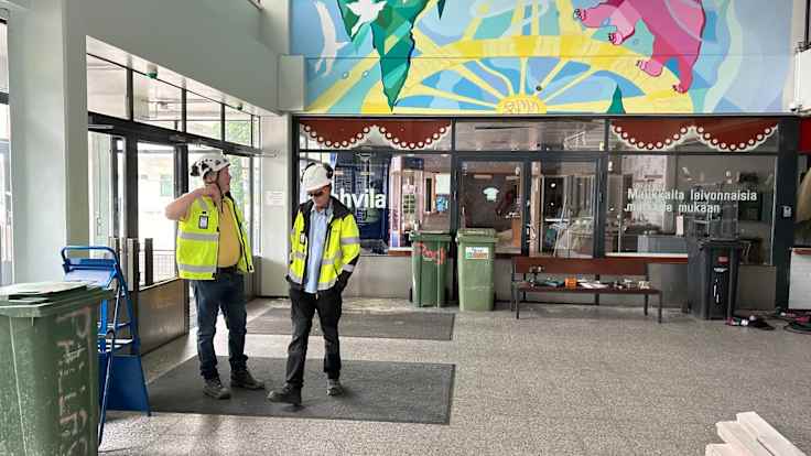 Kaksi miestä työmaa-asuissa seisoo muraalin alla remontin keskellä olevan matkakeskuksen aulassa.