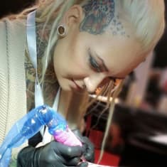 Tatuoija tekee asiakkaalleen tatuointia. Tatuoija on vaaleahiuksinen nainen, jolla on vaalea neuletakki ja tatutointeja rintakehässään, kaulassaan ja ohimollaan.