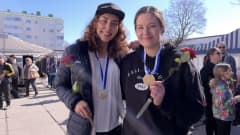 Kaksi hymyilevää naista, joilla kultamitalit kaulassa ja ruusut kädessä. 