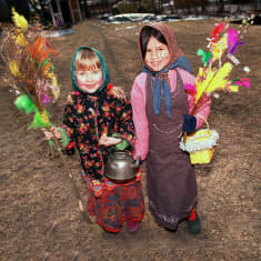 Kaksi noidaksi tai trulliksi pukeutunutta lasta höyhenin koristettuine virpomisoksineen hymyilee ja katsoo kameraan.