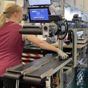 En kvinna står och plockar plastdelar från ett rullande band vid en robot i en fabrik.