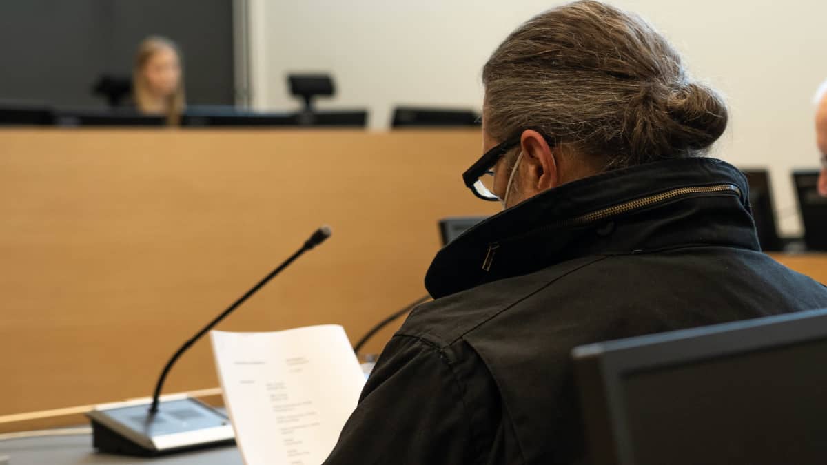 Mustatakkinen mies istuu oikeussalissa. Hänestä näkyy takaapäin kuvattuna hartiat, harmaat hiukset ponihäntänutturalla niskassa, tummasankaiset silmälasit. Mies lukee asiakirjaa. 