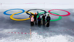 Joukko ihmisiä seisoo isojen jäähän tehtyjen ja värikkäiden olympiarenkaiden edessä. Osa vilkuttaa ylöspäin kameralle.