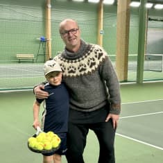 Seitsenvuotias Eemil Koivisto ja Veli Paloheimo seisovat vieretysten tenniskentällä ja katsovat hymyillen kameraan. Eemil pitelee kädessään tennismailaa, jonka verkon päällä on kasa tennispalloja.