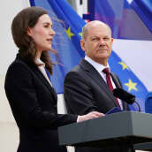 Sanna Marin, Olaf Scholz och magdalena Andersson håller presskonferens bakom var sitt podie i blåsiga förhållanden. I bakgrunden ländernas flaggor.