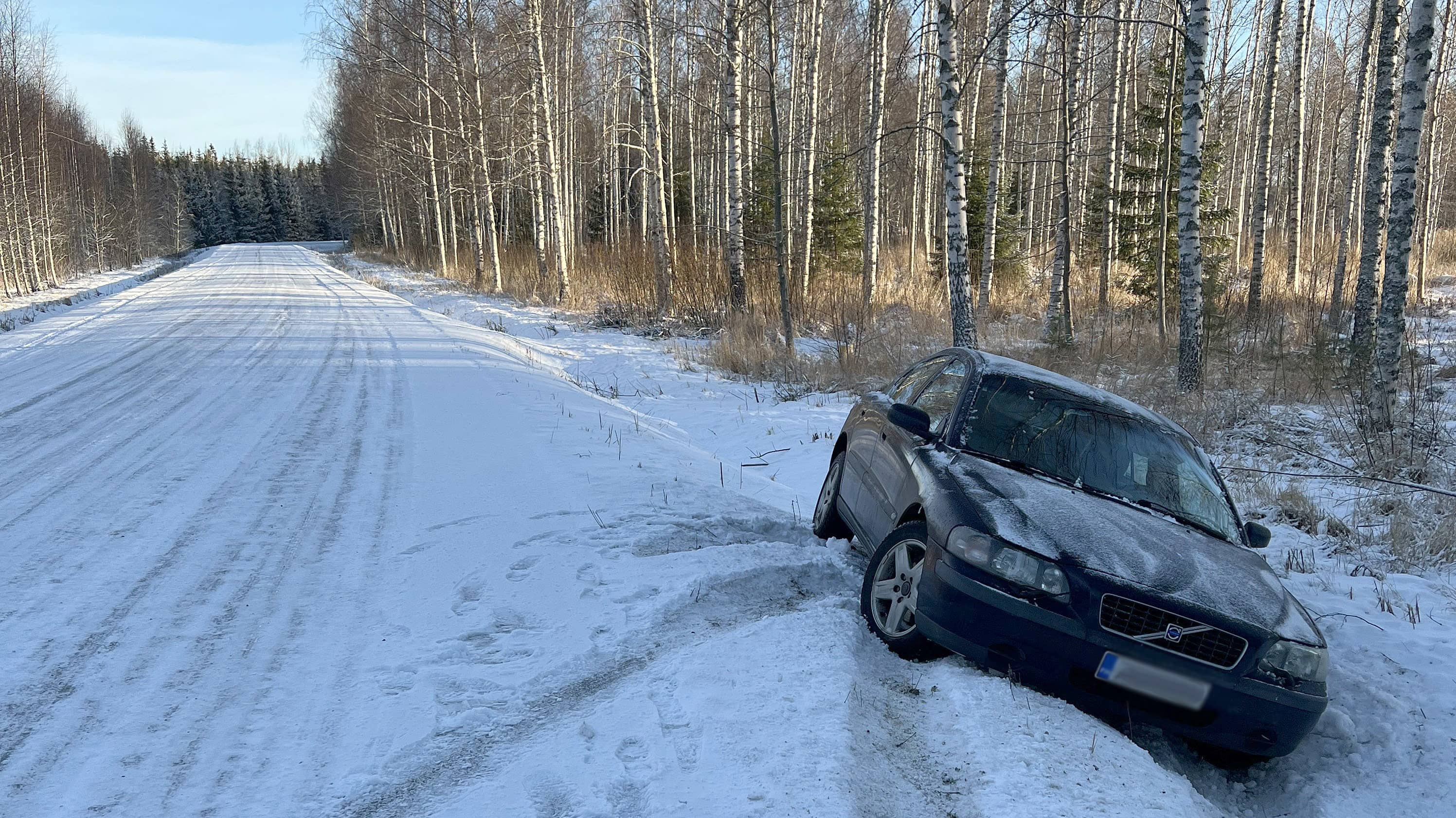 Tummansininen Volvo oikealla puolella tietä ojassa talvella, auton nokka on kohti kameraa, tien ympärillä koivikkoa.