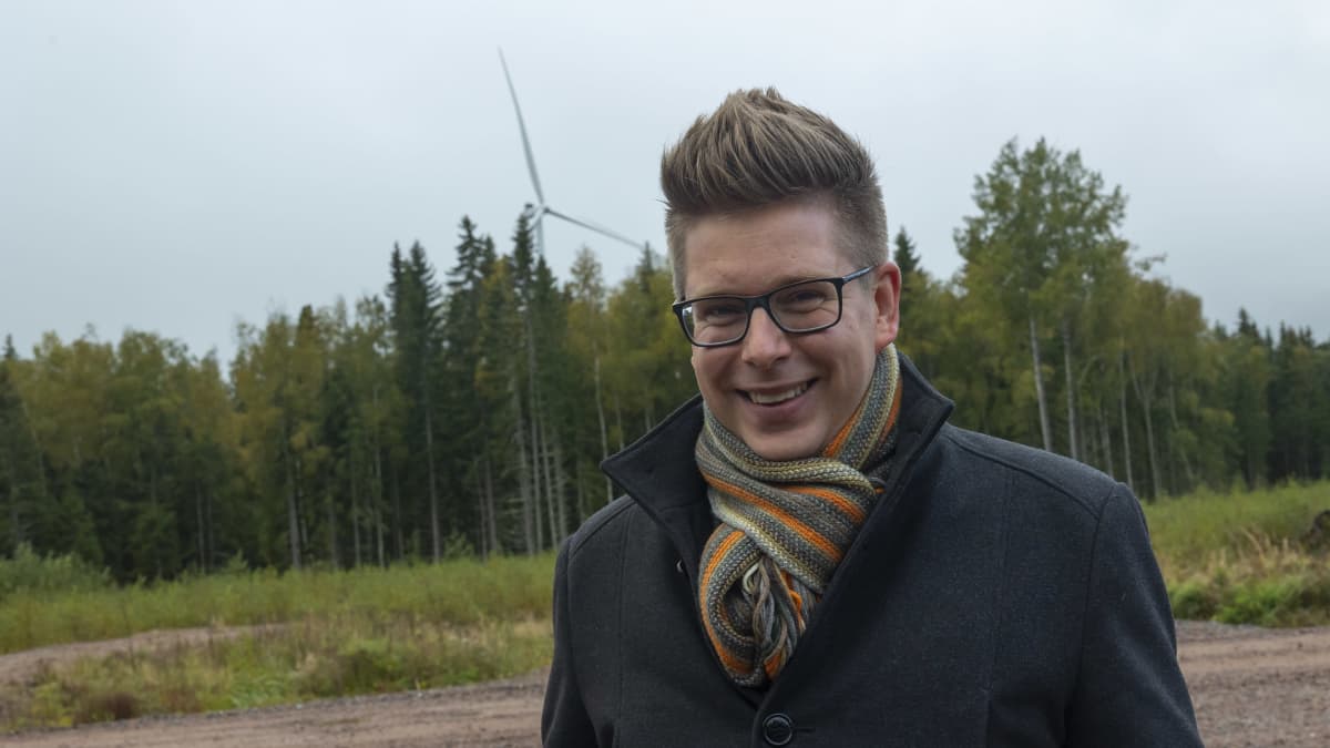 Isojoen kunnanjohtaja Juha Takala hymyilee kameralle. Takana metsässä näkyy tuulivoimalan lavat.