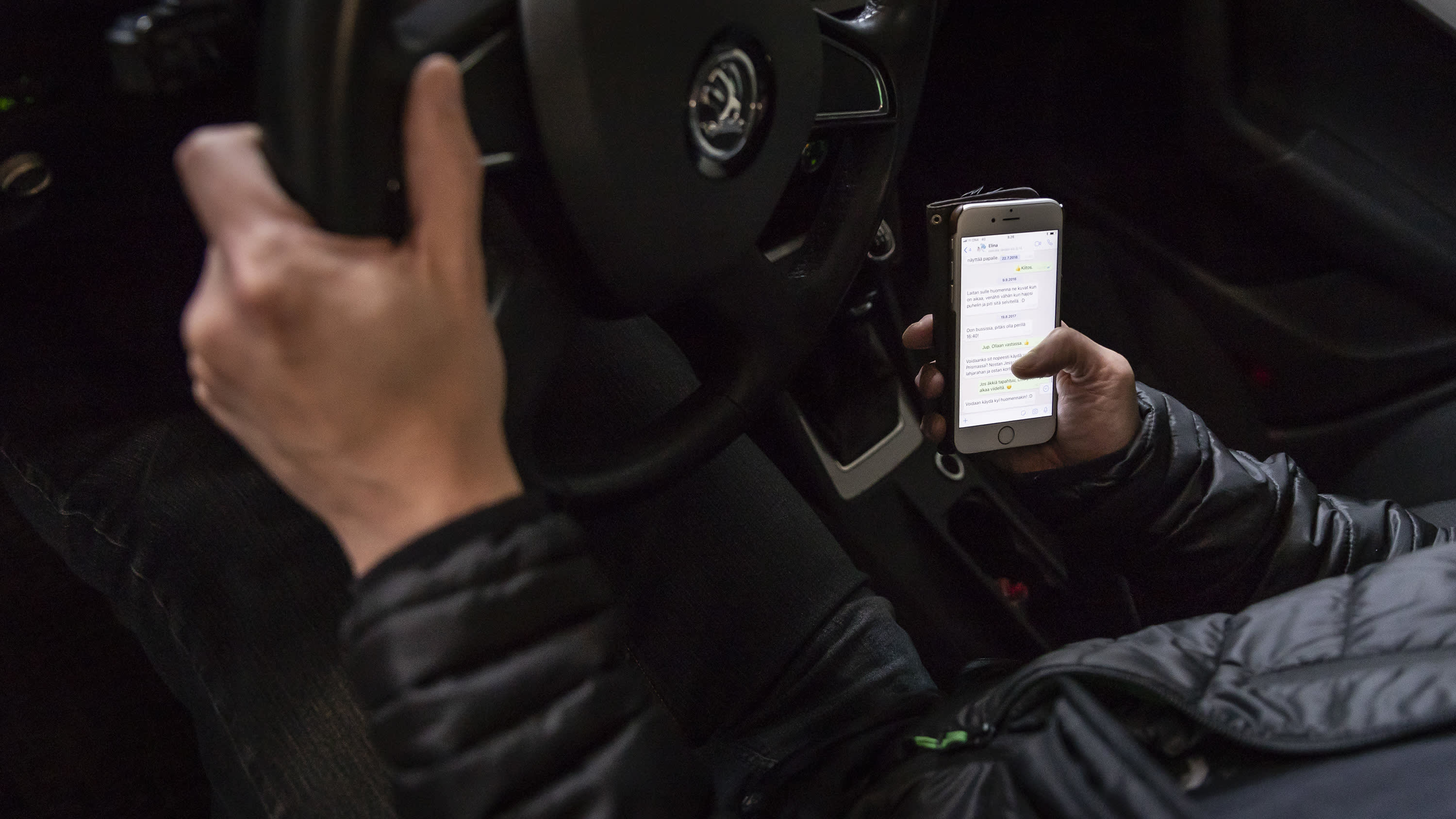Autoilijan toinen käsi on ratilla ja toinen käyttää älypuhelinta. 