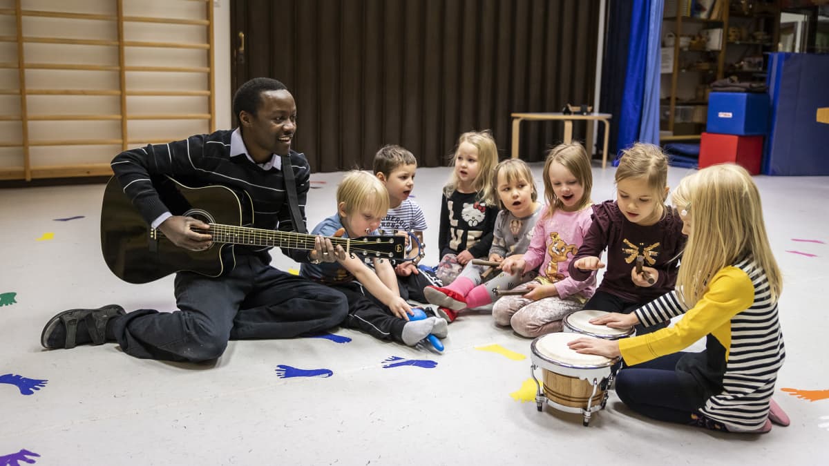 Vasemmalla kitaraa soittava Malloy Nyakwama ja hänen oikealla puolellaan erilaisia instrumentteja soittavia lapsia. 