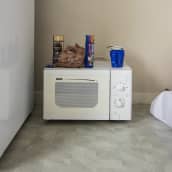 Pakolaisnaisten huoneen lattialla on mikroaaltouuni, jonka päällä on keksipakkauksia, pikakahviajauhepurkki ja kaksi mukia.  Mikron vieressä siintävät jääkaappi ja lattialle asetettu patja. 