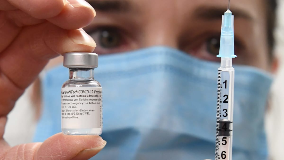 I bakgrunden syns ett ansikte med munskydd. Kameran fokuserar på en liten glasflaska med coronavaccin och en spruta personen håller i.