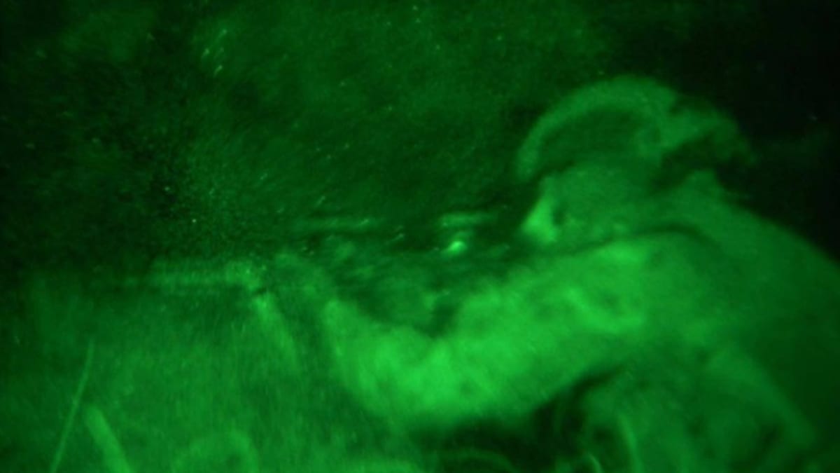 Kuvakaappauksessa sotilas tähtää kiväärillä pimeässä.