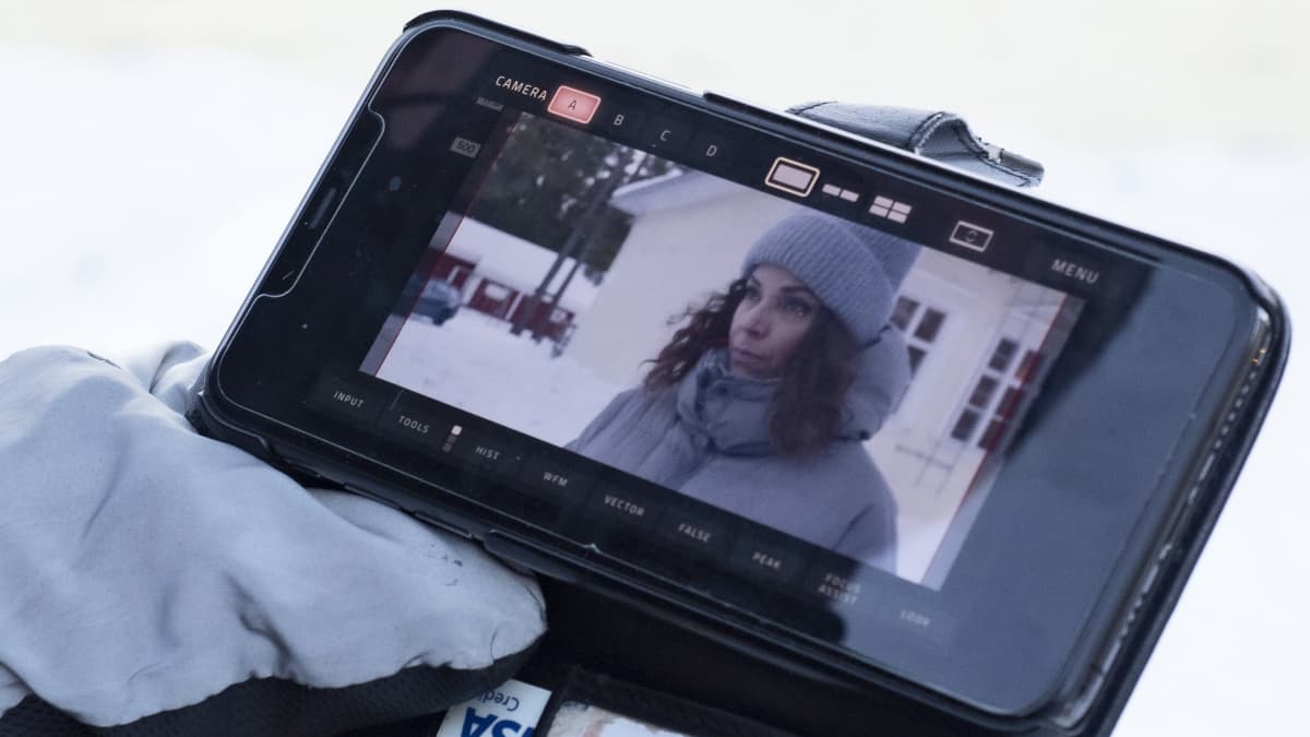 Kädessä kännykkä, jossa näkyy kuva talvivaatteisiin pukeutuneesta Amira Khalifasta näyttelemässä kohtausta tv-sarjassa.
