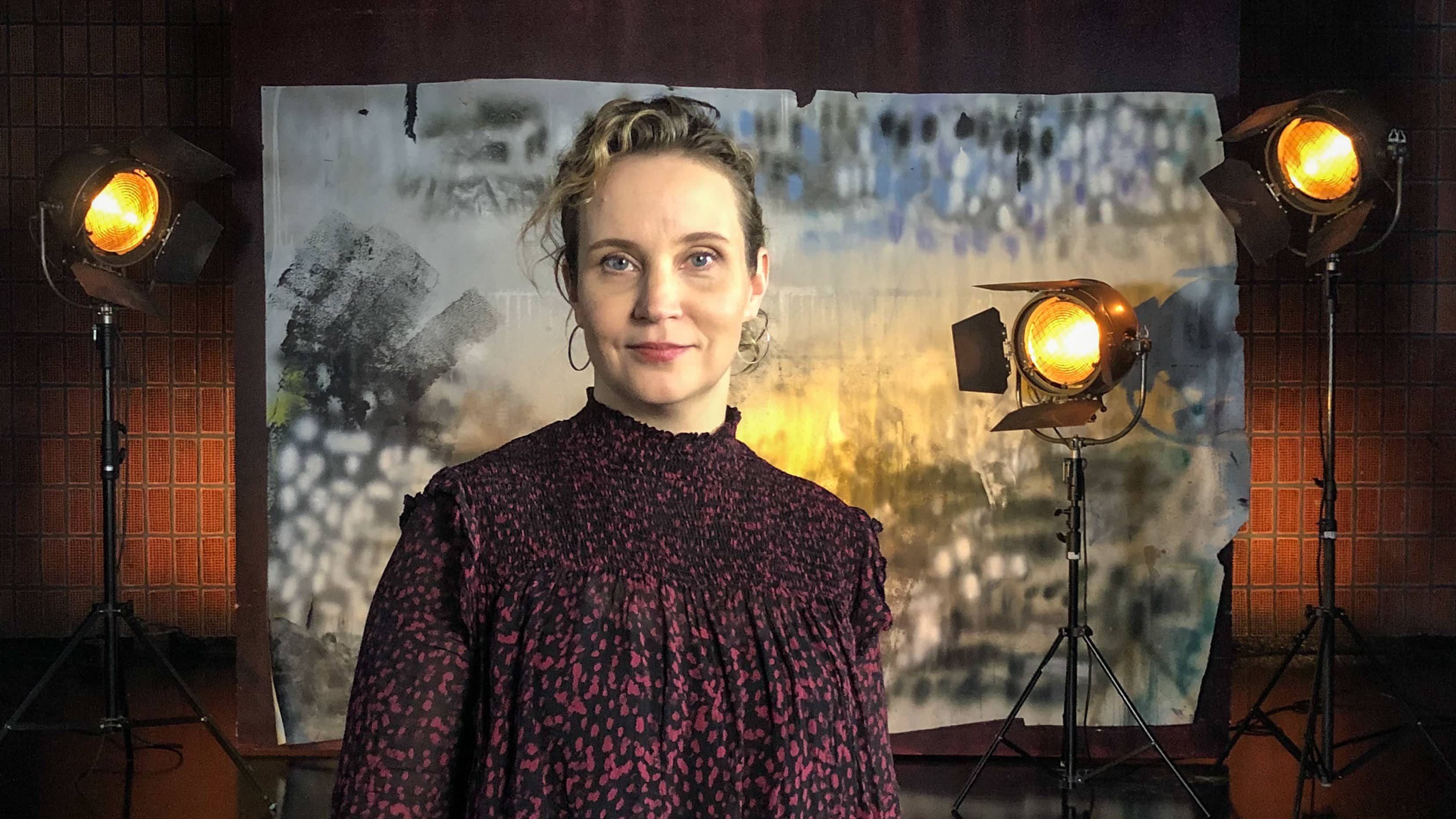 Kirsikka Saari seisoo studiossa taustalavasteen ja studiolamppujen edessä. Kuva otettu lyhytvideosarjaan Me elokuvantekijät.