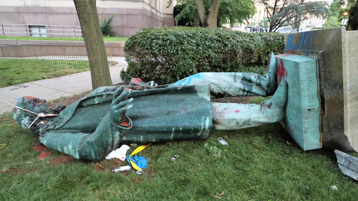 Albert Piken kaadettu patsas makaa maassa nurmikolla, patsaan kaulassa on köysi. Patsasta on töhritty punavärillä.