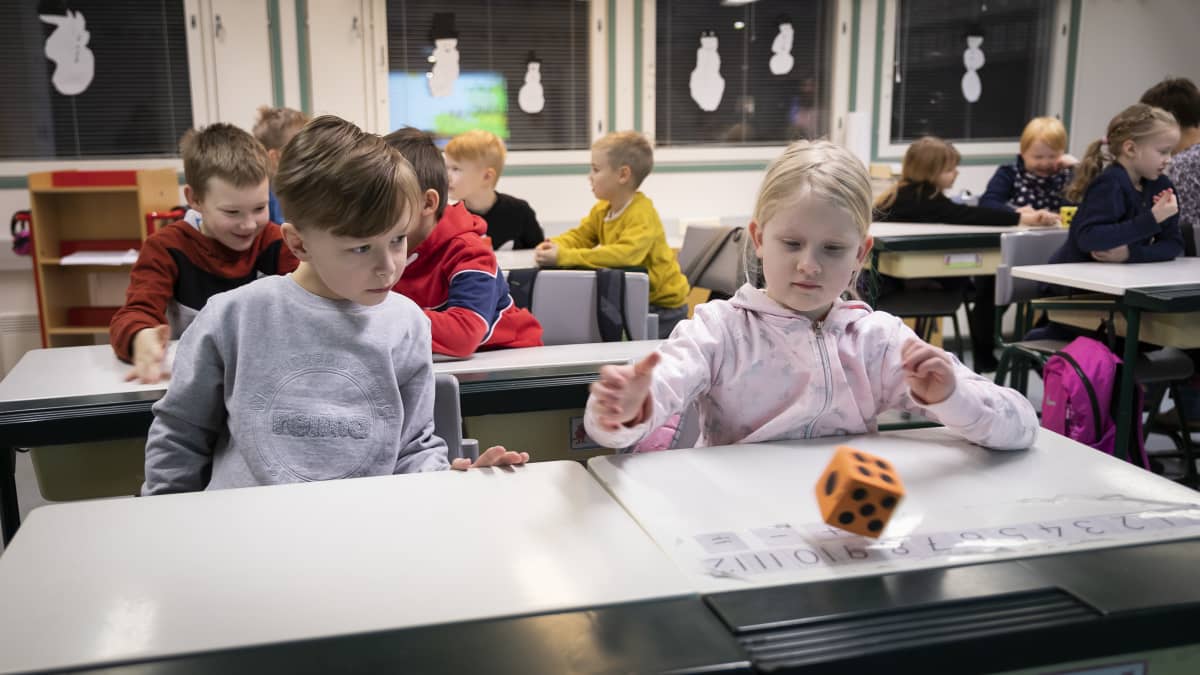 Lapsia istumassa pulpeteissaan koululuokassa. Oikealla lapsi heittää oranssia noppaa. Vasemmalla oleva lapsi tarkkailee vierestä. 