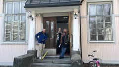 Kolme ihmistä, Adam Huuva, Katariina Salo ja Merja Ruth seisovat talon oven ulkopuolella. Oven edessä on puiset pylväät ja kiviportaat ja ikkunat ovat pikkuruutuiset.