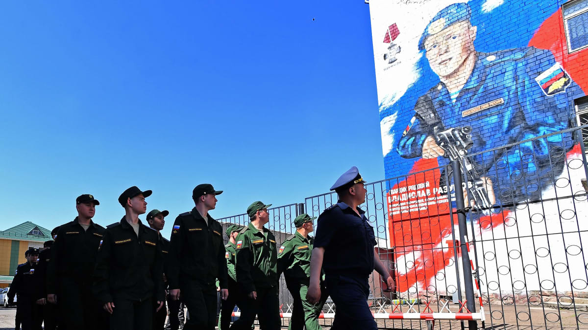 Joukko vihreissä univormuissa olevia sotilaita marssii aidan viertä ohi suuren seinämaalauksen, joka esittää nuorta siniseen univormuun pukeutunutta sotilasta rynnäkkökivääri kädessään.