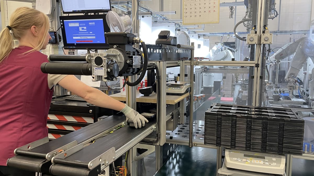 En kvinna står och plockar plastdelar från ett rullande band vid en robot i en fabrik.