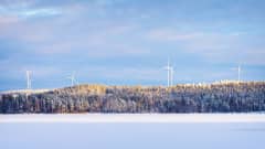 Tuulivoimaloita horisontissa kukkuloiden takana. Etualalla jäätynyt järvi, jonka päällä on lunta.
