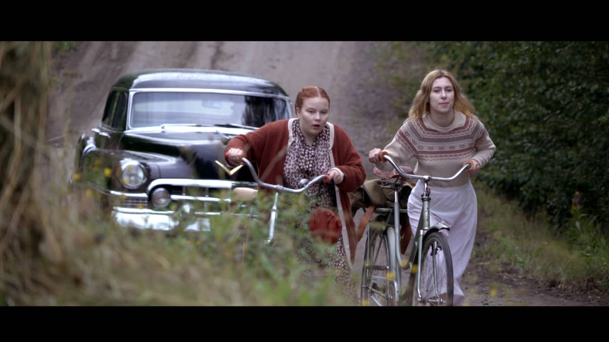 Elokuvan Kyllikki työntää polkupyörää ystävänsä kanssa. Taustalla auto.