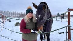 Nuori vaaleahiuksinen nainen seisoo hevosaitauksen edessä, maassa on lunta. Aitauksen kalterien yli kurottaa iso ruskea hevonen, joka haistelee naisen kämmentä.