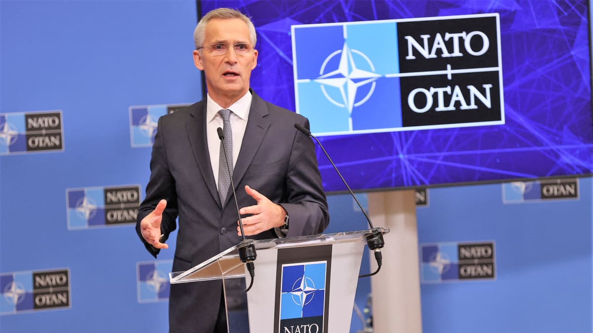 Stoltenberg puhuu. Hänen edessään on pieni puhujanpönttö, jonka päällä sojittaa kaksi mikrofonia. Stoltenbergillä on tummanharmaa puku ja vaaleanharmaa kravatti. Taustalla näkyy Naton logo ja tekstit NATO - OTAN.