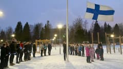 Lipputangon ympärillä on isoja ja pieniä koululaisia. Yksi isoista koululaisista nostaa Suomen lippua salkoon ja tuuli on lennättänyt lipun kauas tangosta.
