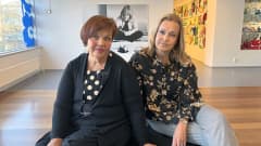 Helena Lindstén ja Hanna Mamia-Walther istuvat Riihimäen taidemuseon penkillä, taustalla heidän välissään valokuvasuurennos Maija Isolasta.
