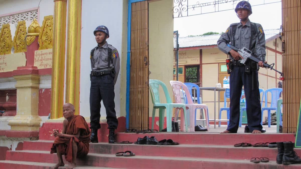 Punaiseen kaapuun pukeutunut munkki istuu portailla. Hänen takanaan sotilaita, joista toinen pitelee rynnäkkökivääriä.