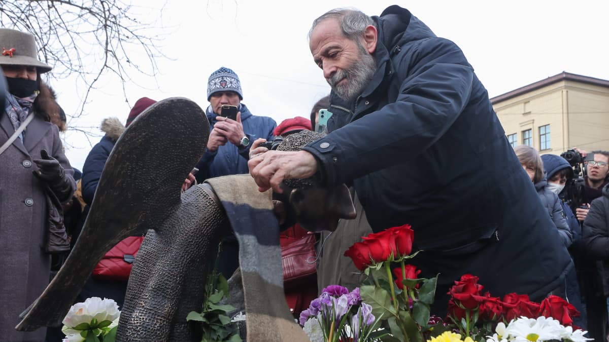 Parrakas mies Boris Vishnevski jättää kukkia enkelipatsaalle, ympärillä ihmisiä ja kukkia