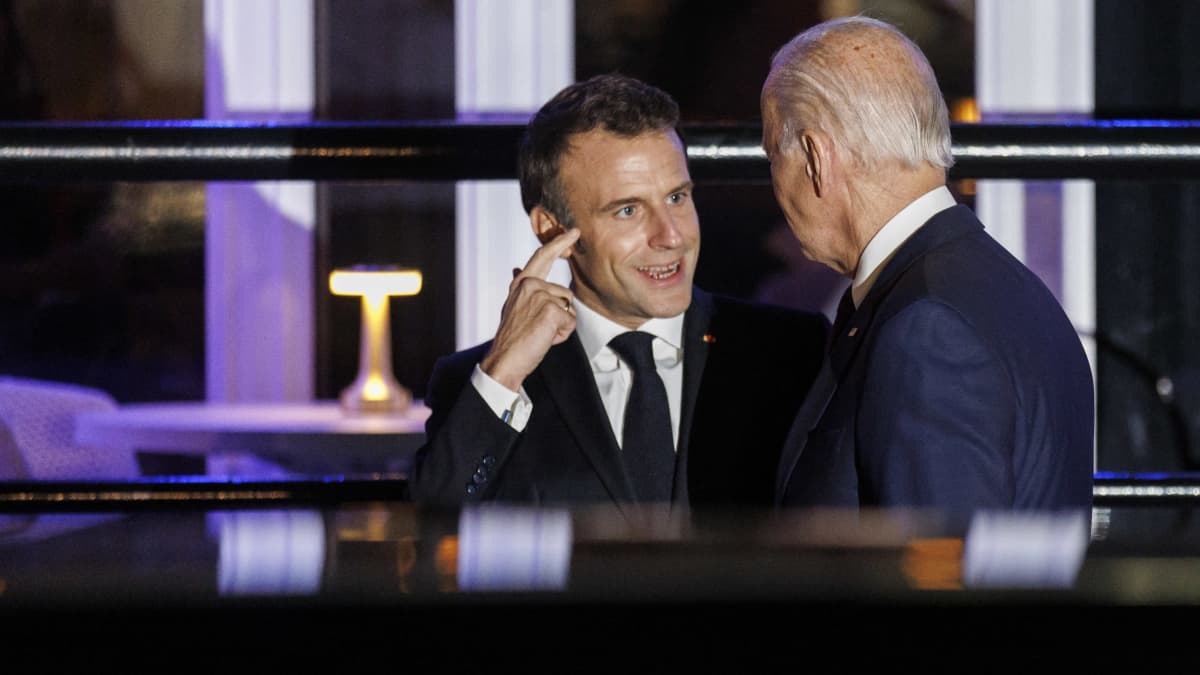 Macron osoittaa ohimoaan etusormellaan ravintolan edessä, Biden selin kuuntelee, mitä tämä sanoo.