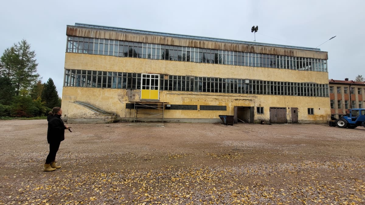Keltainen, rapistunut suorakaiteen mallinen tehdasrakennus, jota kiertävät kahdessa kerroksessa ikkunarivistöt. Etualalla seisoo nainen selin.