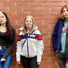 Parikymppiset tummahiuksinen tyttö, jolla jalassa farkut ja ulkoilupusakkaan pukeutunut vaaleahiuksinen tyttö sekä polkkatukkainen siniseen takkiin ja mustiin farkkuihin pukeutunut poika seivat punatiiliseinän edessä.  