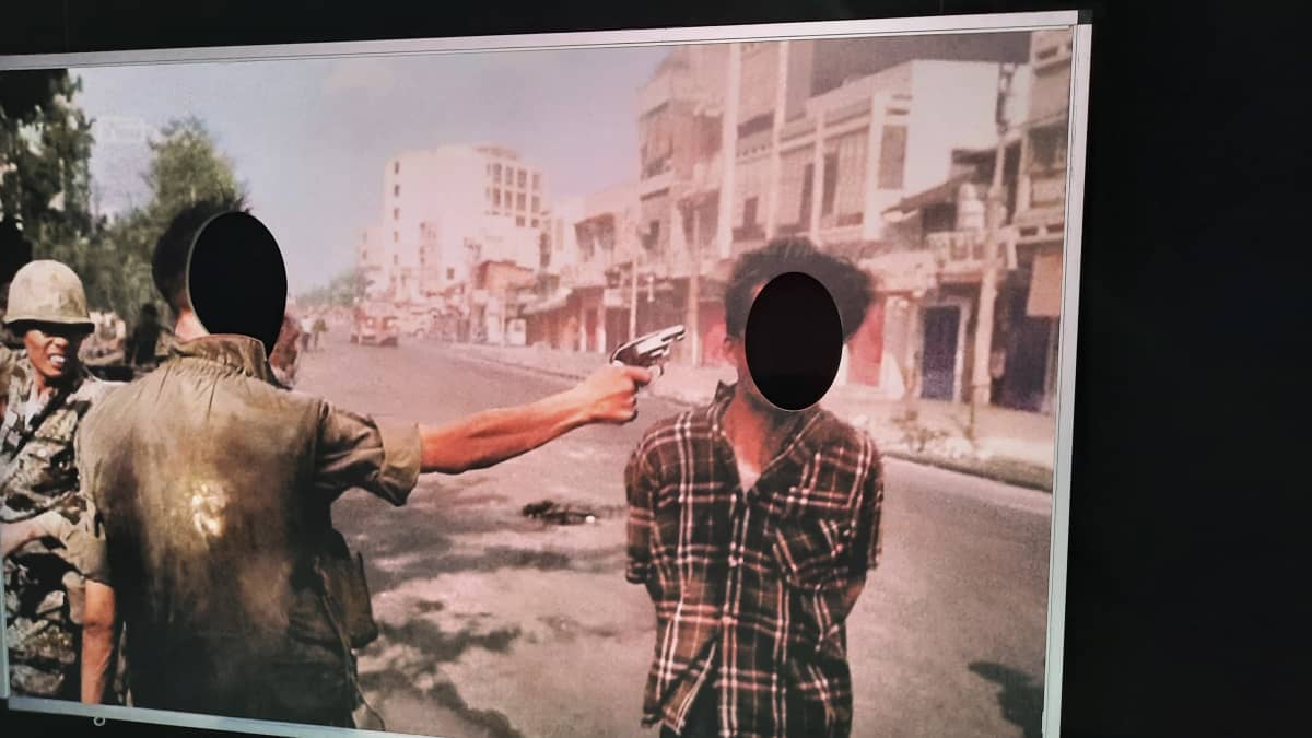 Kuvassa pahviseinäke Eddie Adamsin kuuluisasta valokuvasta Vietnamin sodasta, jossa toinen ihminen ampuu toista päähän. Naamojen kohdalle on tehty reijät, joista voi sujauttaa omat kasvot kuvan hamojen kasvoiksi.