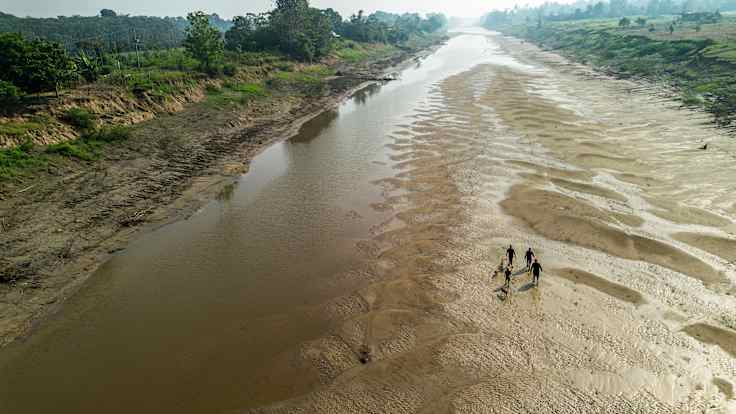 Aerial view of a mostly-dry riverbed, with small silhouettes of four people and a dog walking on the sand with vegetation in the riverbanks.  