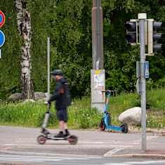 A blurry image of a man wearing a helmet, black jacket and shorts on an e-scooter with another parked behind him and greenery in the background. 