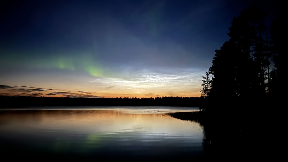 Auringonlaskun jälkeen järven rannalla kuvattu taivasta jolla valaisevia yöpilviä ja hennot revontulet.